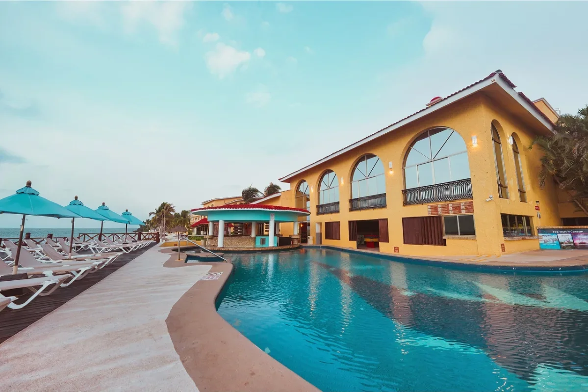 A resort with a swimming pool and yellow building with large arched windows, lounge chairs with blue umbrellas along the poolside, and the ocean in the background under a partly cloudy sky.