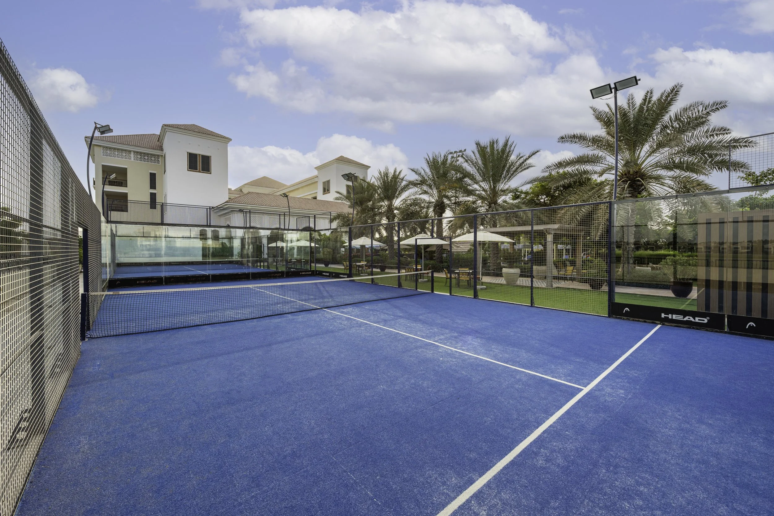 Outdoor blue tennis court surrounded by a high fence, with palm trees, umbrellas, and residential buildings in the background, under a partly cloudy sky.