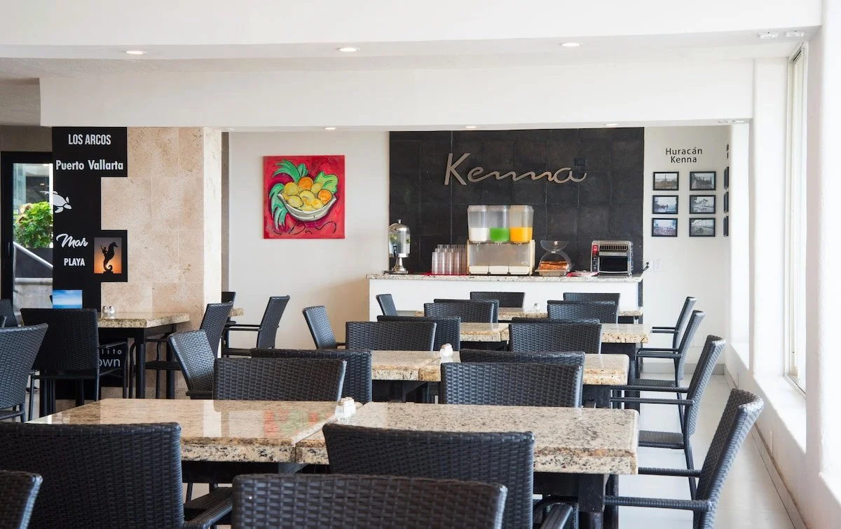Empty dining area with black wicker chairs and granite tables, water dispenser, juice dispenser, small over toaster oven, framed wall art, and a black sign with white text in Spanish on the left.