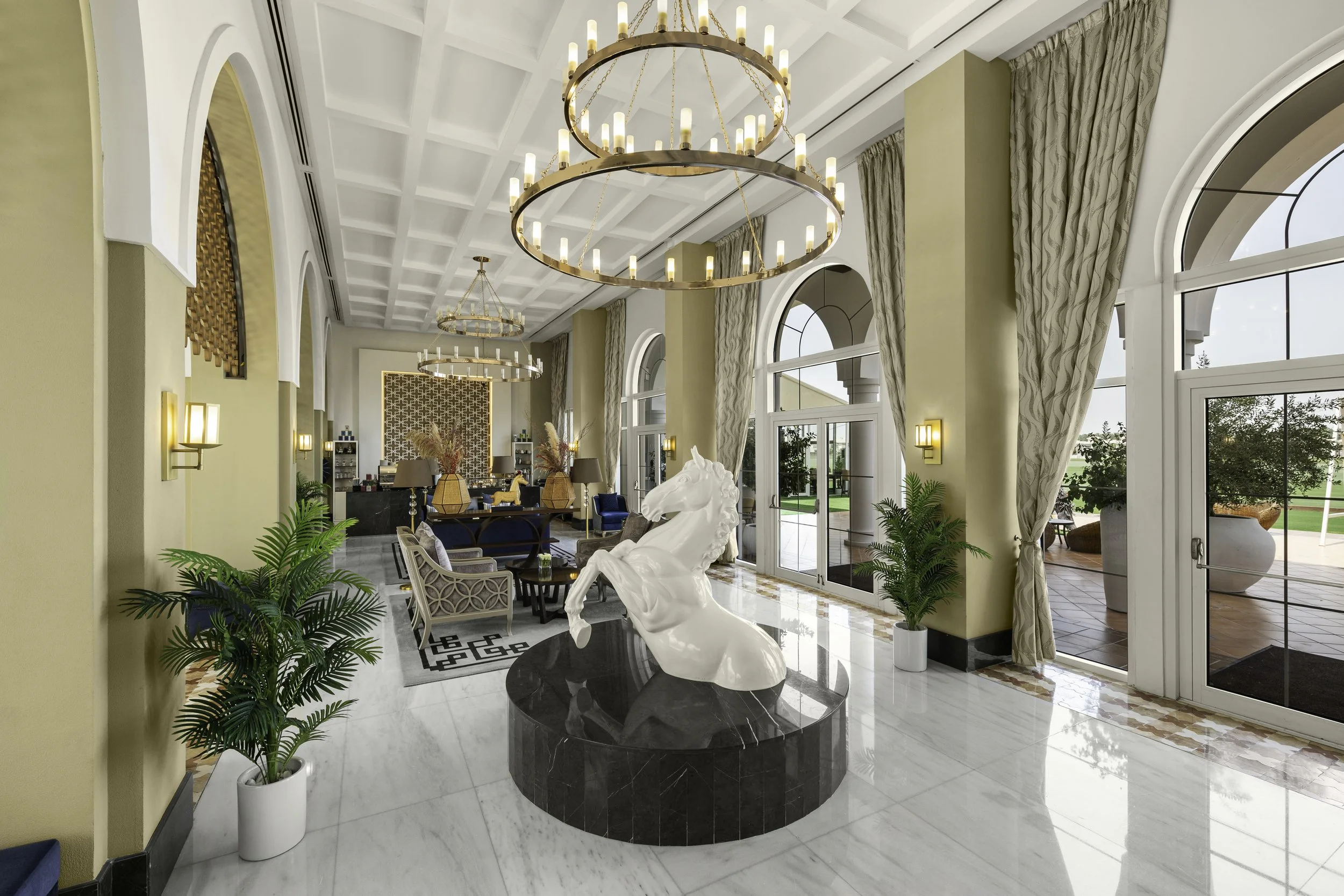 Luxury interior lobby with large windows, hanging chandeliers, a white horse sculpture, and elegant furniture.