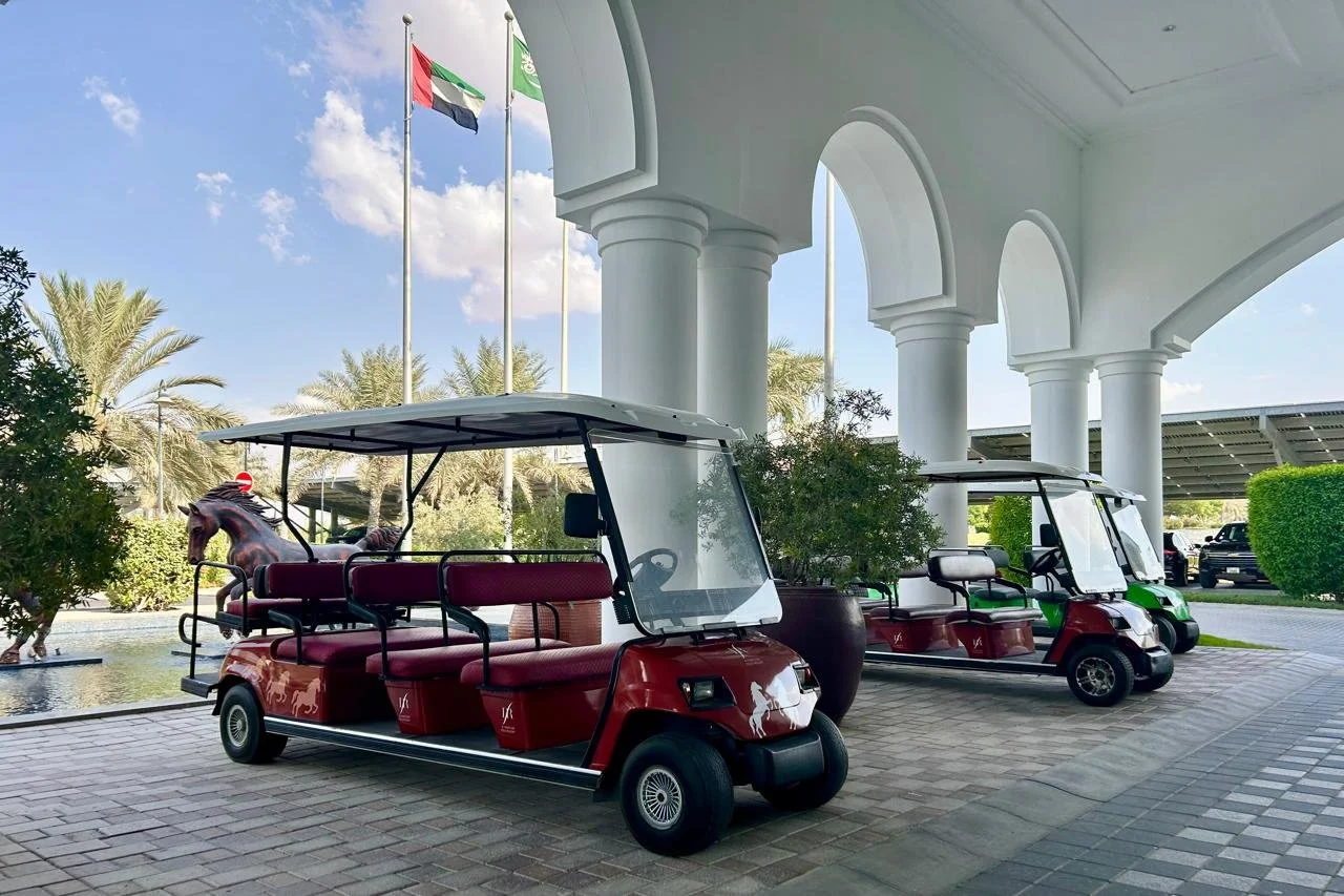 Two red golf carts parked under a white archway, with palm trees and flags in the background.