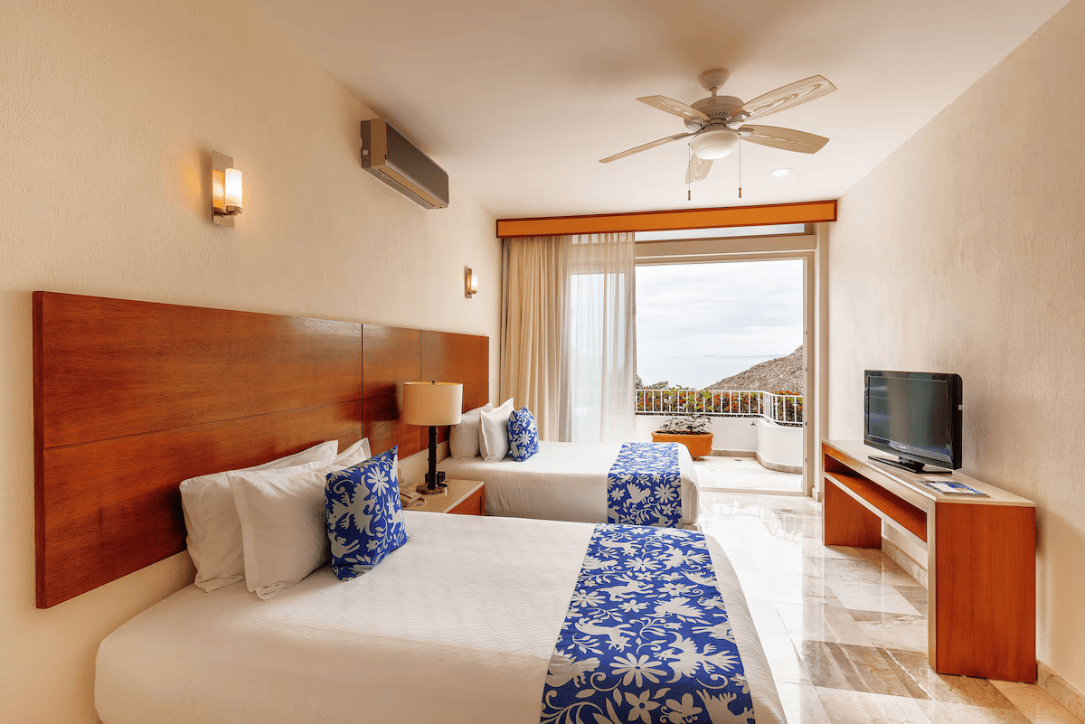 Room with two beds, wooden headboards, blue and white patterned pillows and bed runners, a window with a view of mountains and sky, a ceiling fan, a TV on a wooden stand, and a doorway leading to a balcony with a potted plant.