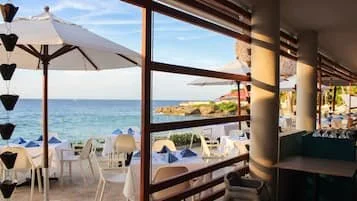 Scene of an outdoor restaurant or cafe with umbrellas, tables, and chairs overlooking the ocean and sky.