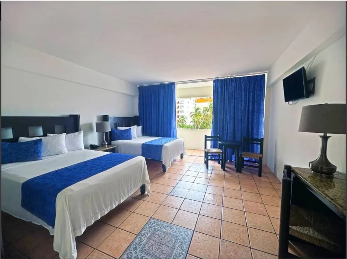 Hotel room with two double beds, blue bedding, nightstand with lamps between beds, window with blue curtains, two chairs, small table, wall-mounted TV, and tiled floor.