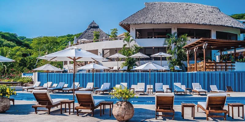 Resort pool area with lounge chairs, umbrellas, and potted plants, with a thatched-roof building and lush green trees in the background.
