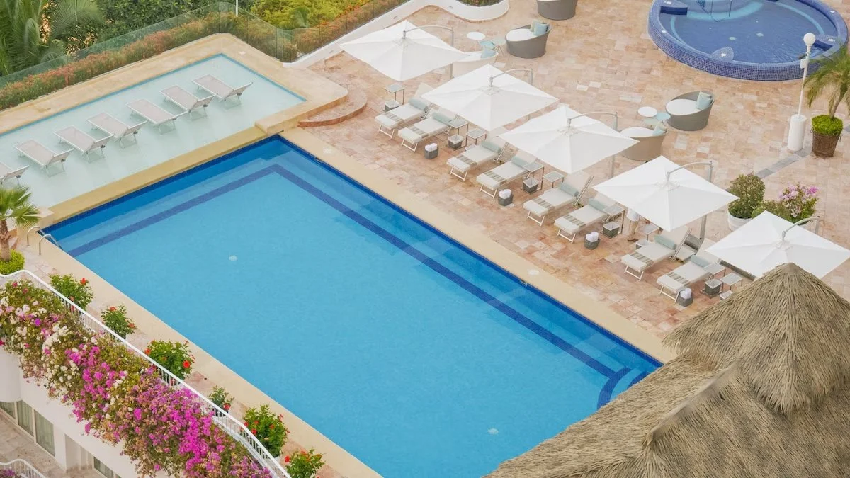 Aerial view of a hotel pool area with lounge chairs, umbrellas, a hot tub, and colorful flowers along the railing.