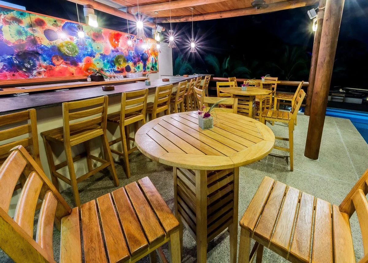 Empty outdoor bar or restaurant seating area at night with wooden tables and chairs, colorful abstract wall art, hanging lights, and a view of palm trees and a pool in the background.