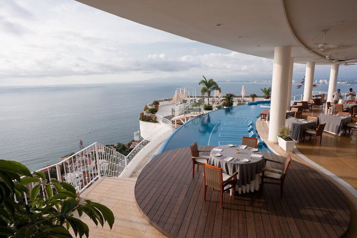 An outdoor restaurant terrace with round tables covered in gray tablecloths, set with plates and glasses, overlooking a curved swimming pool and the ocean with a distant shoreline and cloudy sky.