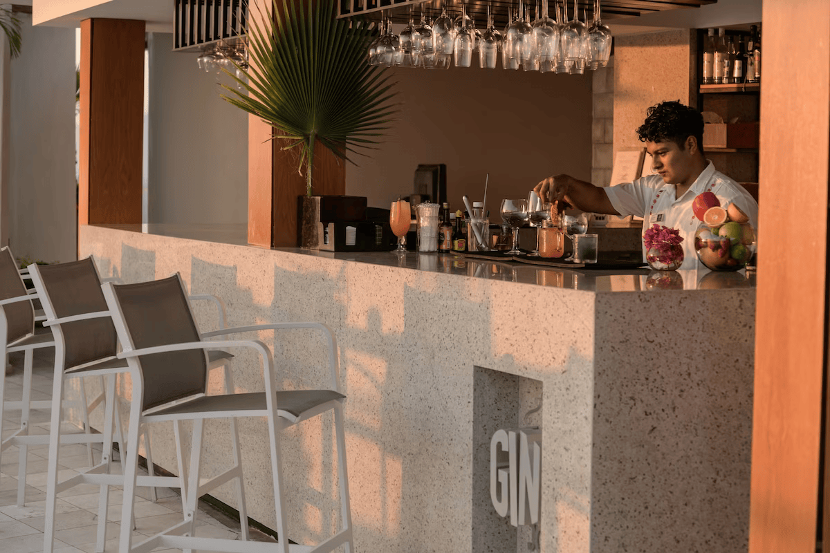 A bartender preparing drinks at a modern bar with hanging wine glasses, a large palm leaf, and bowls of fruit. Four chairs are lined up in front of the bar.