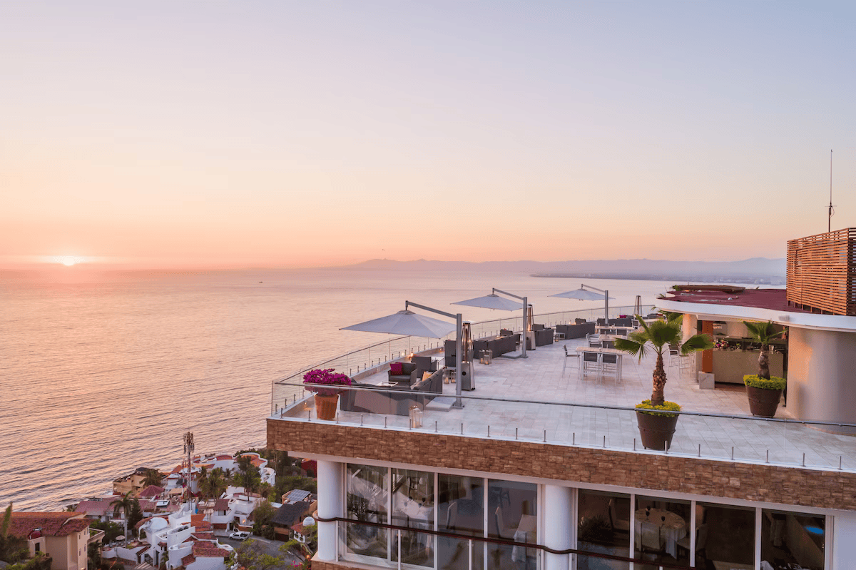Luxury rooftop terrace overlooking the ocean at sunset with seating, umbrellas, potted plants, and a view of the coastal town below.