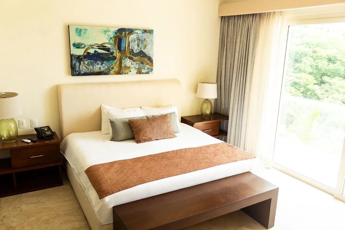 Hotel bedroom with a large bed, nightstands with lamps, abstract wall art, and sliding glass door with curtains showing greenery outside.