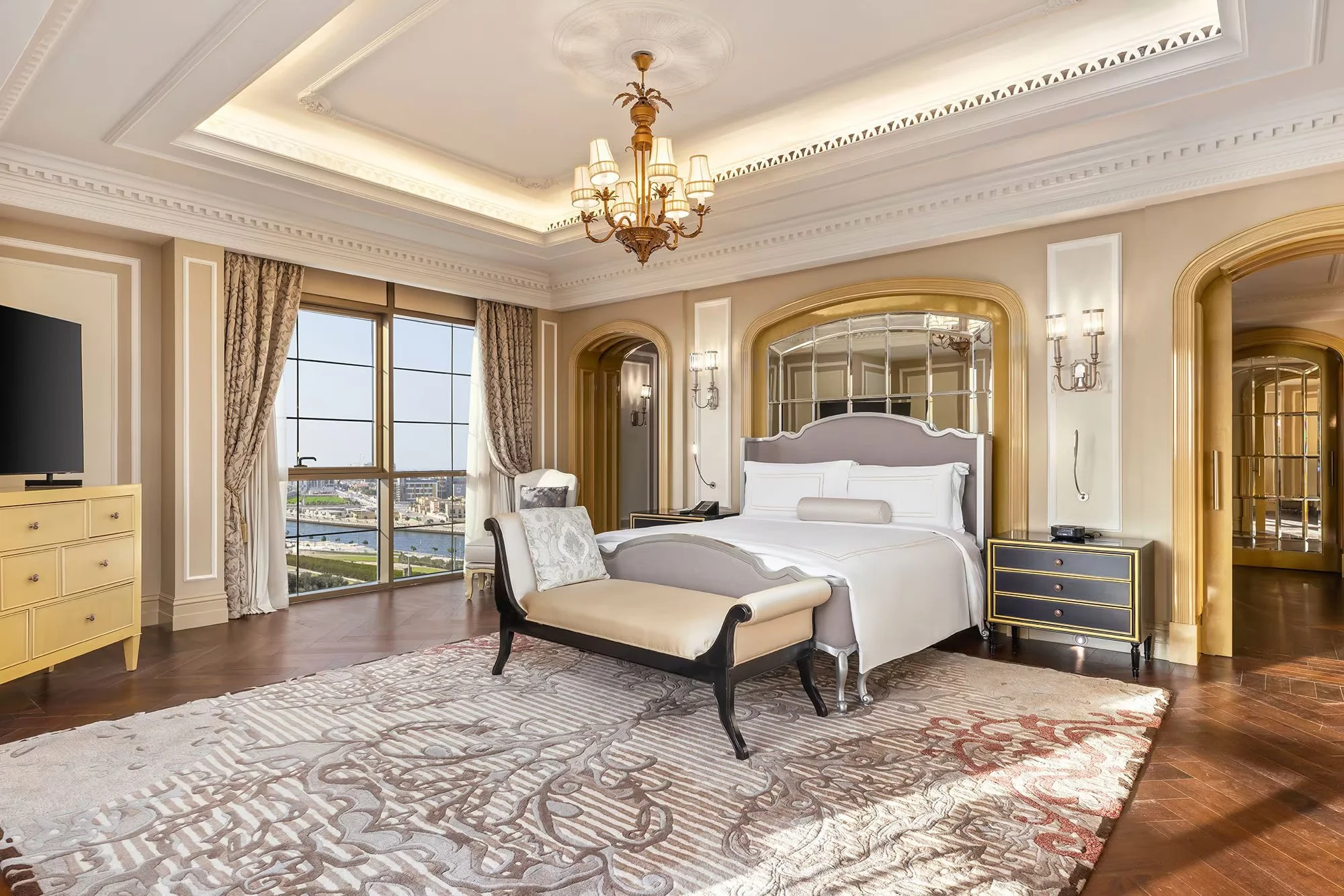 A luxurious hotel bedroom with a large bed, chandelier, and large windows offering a city view.