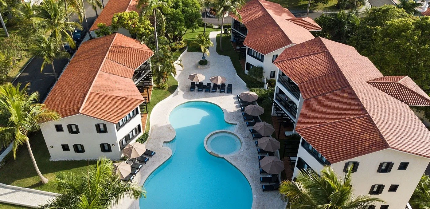 An aerial view of a resort complex with a large swimming pool surrounded by lounge chairs and umbrellas, white multi-story buildings with red tile roofs, and lush green trees.