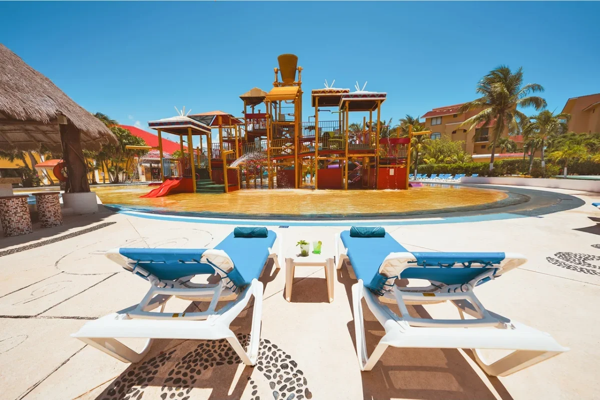 Poolside view with two blue lounge chairs and a small side table in front of a colorful water playground with slides and water features, surrounded by palm trees and residential buildings under a clear blue sky.