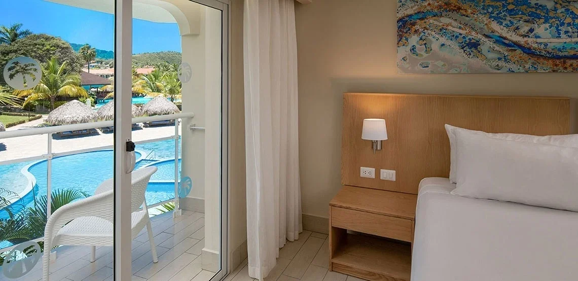 Hotel room with a balcony overlooking a pool and tropical landscaping.