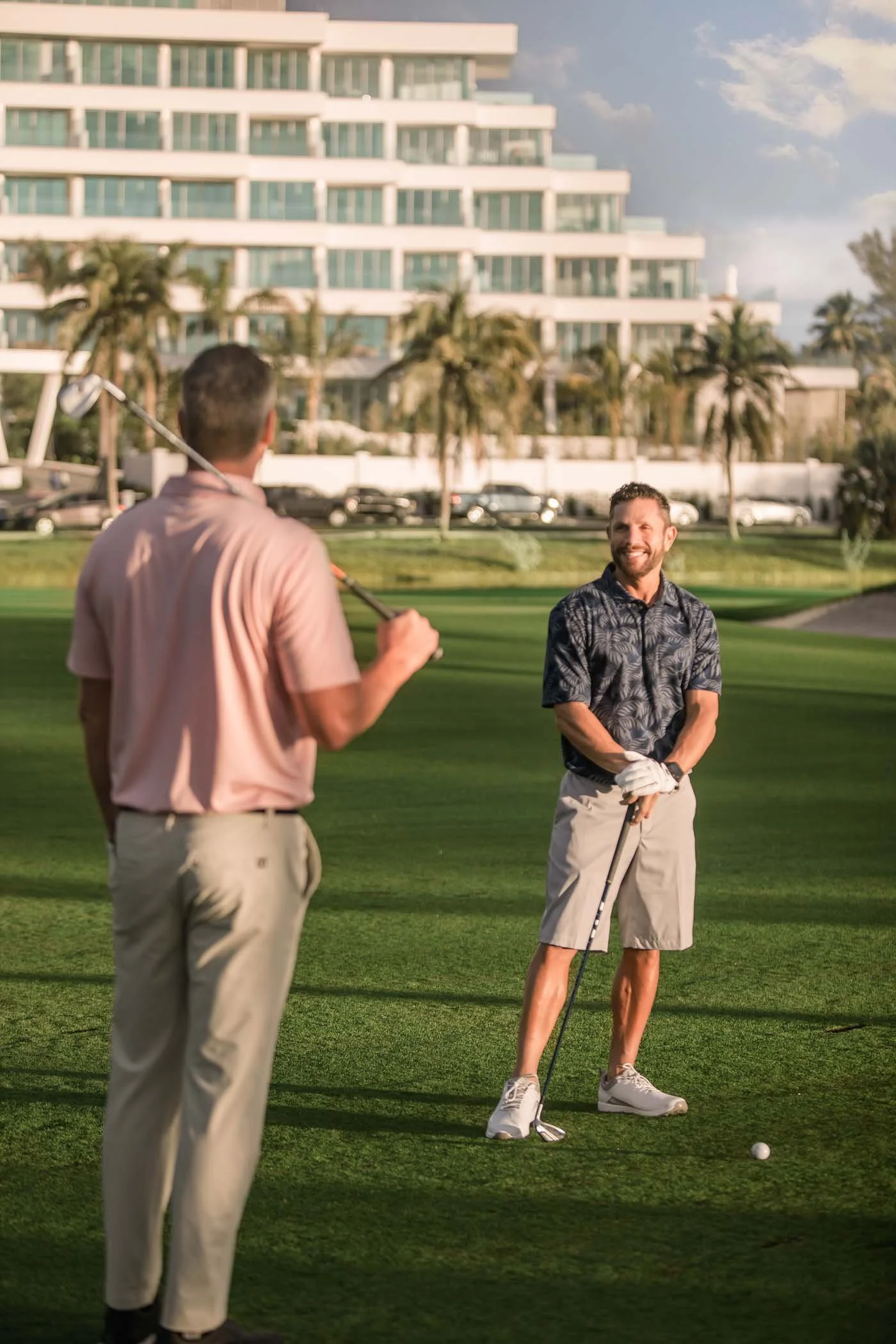 Two men playing golf on a lush green golf course with a large modern building and palm trees in the background, under a partly cloudy sky.