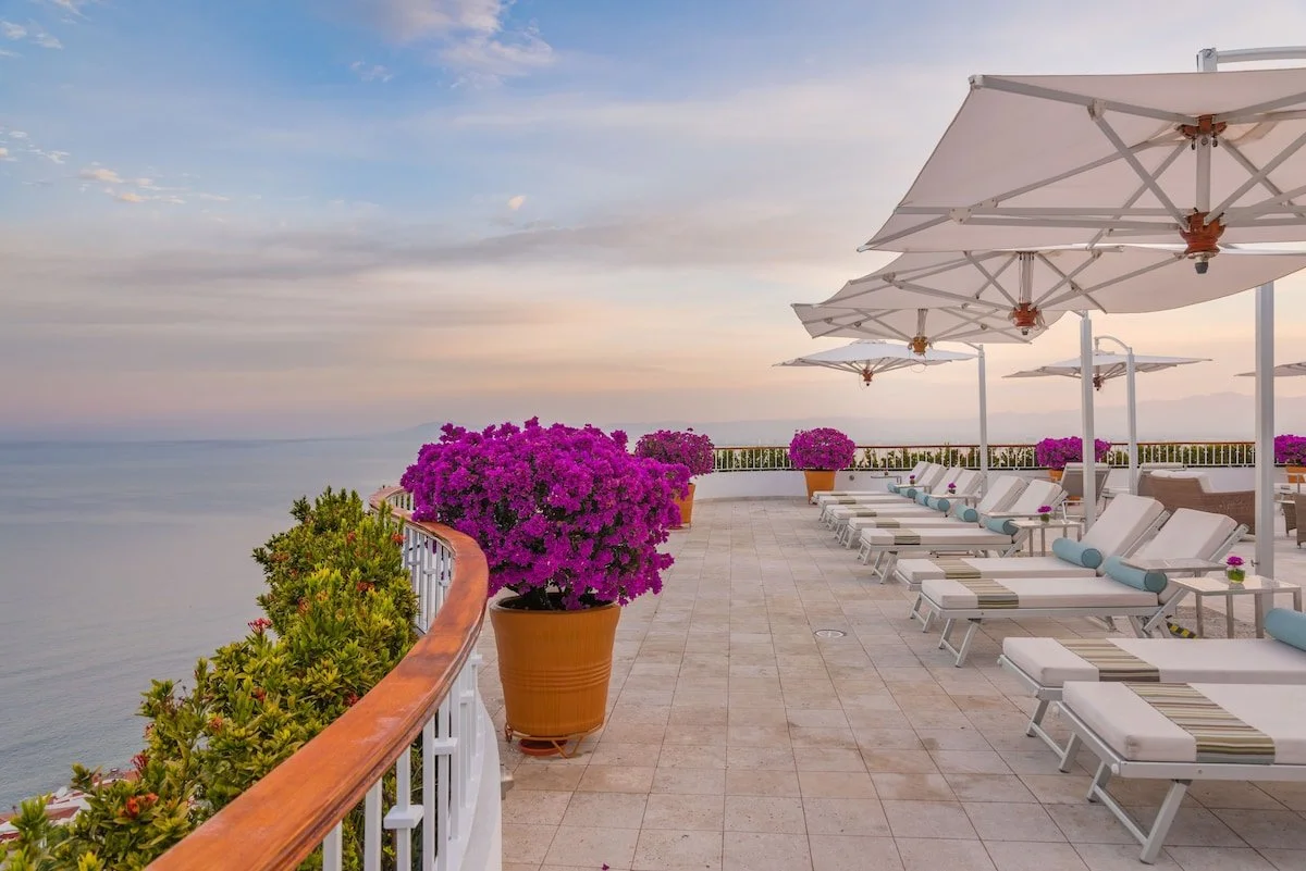 Beachfront terrace with white lounge chairs, large white umbrellas, pink flowering plants in pots, and a view of the ocean during sunset.