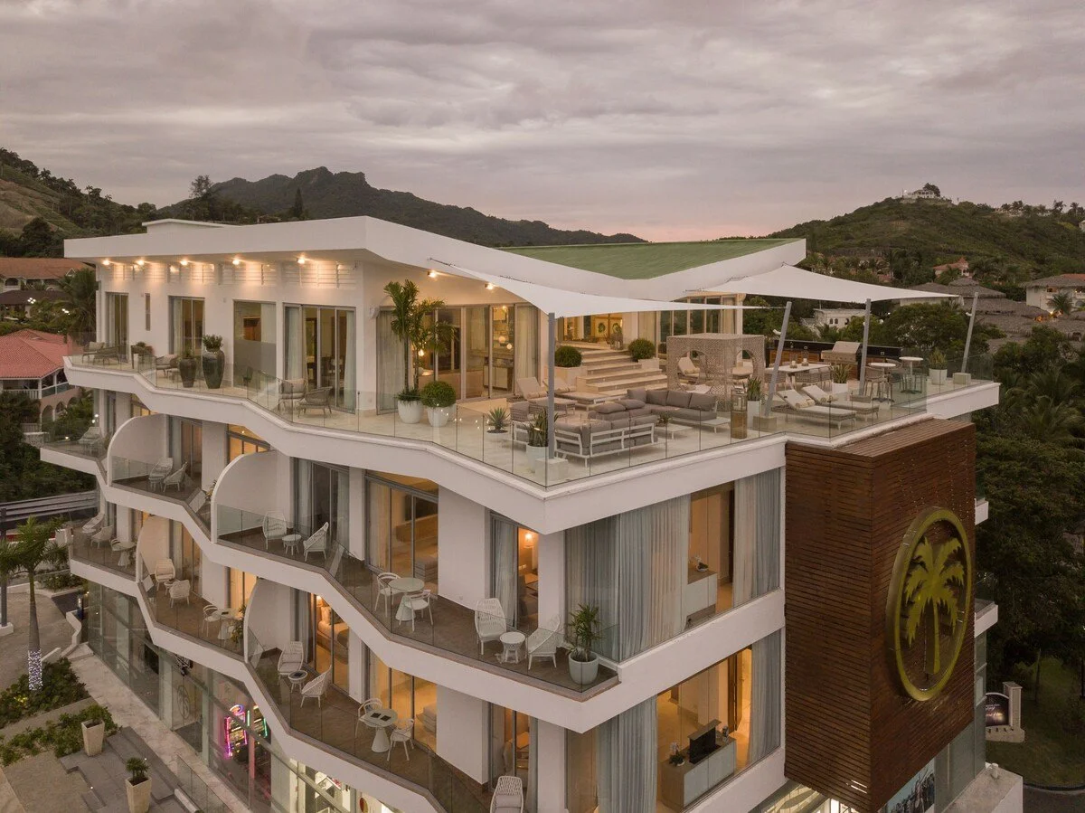 A modern multi-story luxury building with spacious rooftop terrace and outdoor seating, surrounded by hillside landscape at dusk.