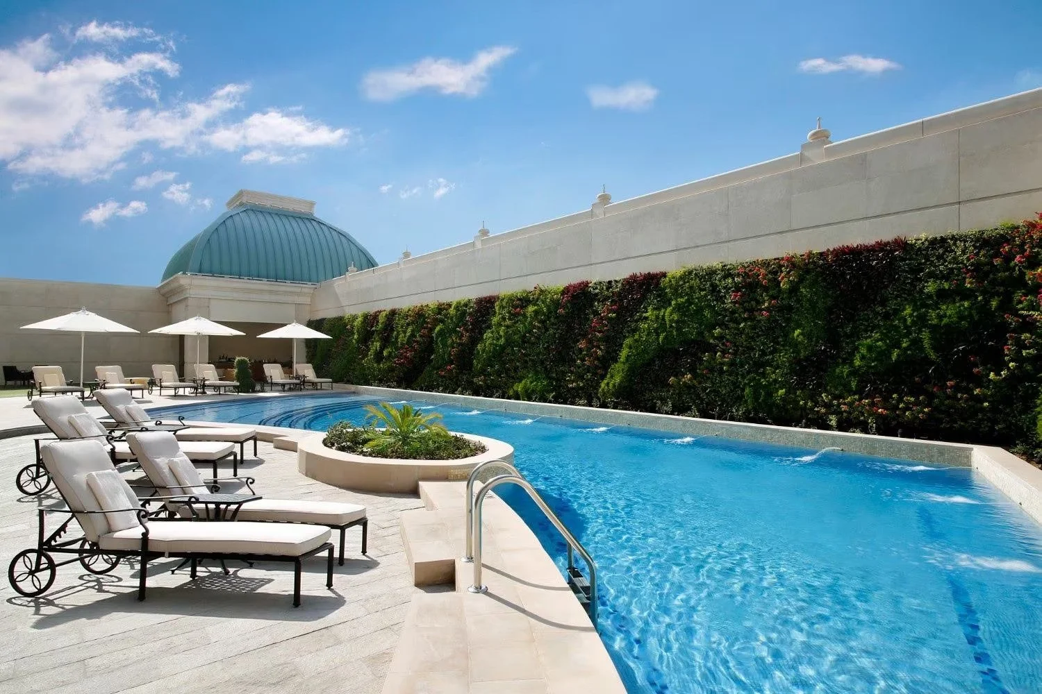 Luxury rooftop pool area with white lounge chairs, umbrellas, a small circular garden with greenery, and a building with a green dome in the background under a partly cloudy blue sky.
