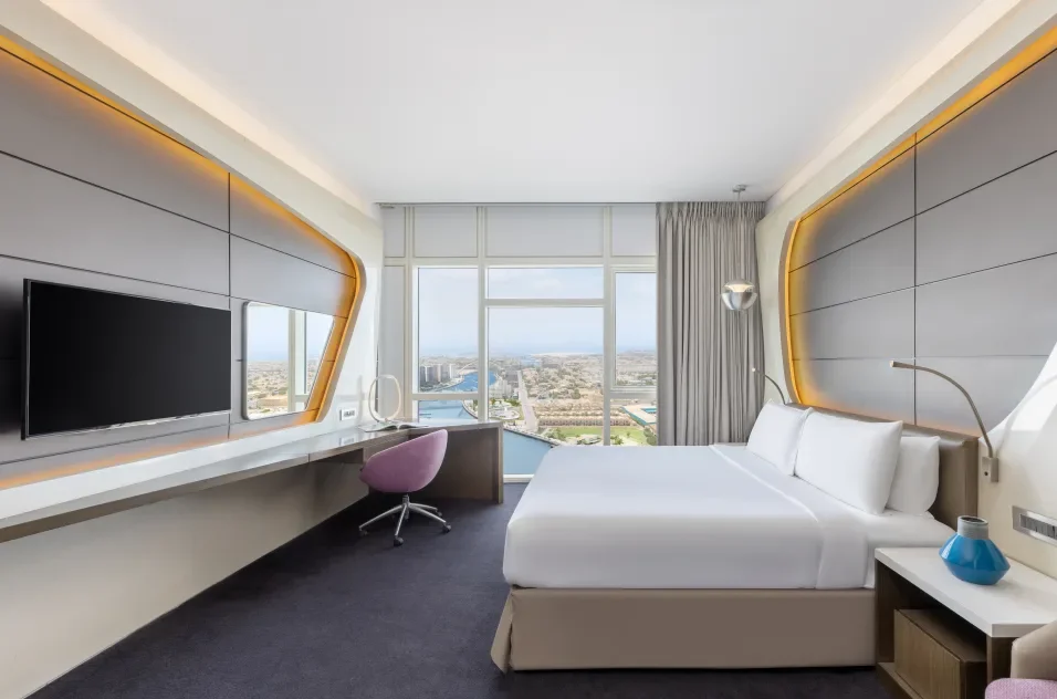 Modern hotel room with a large window offering a city view, a white bed with pillows, a purple chair at a desk, a flat-screen TV, and contemporary decor with grey and white color schemes.