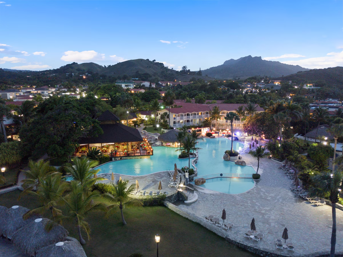 A resort with an outdoor swimming pool and surrounding lounge chairs, umbrellas, palm trees, and a bar with lighting, set against hills and mountains in the background during twilight.