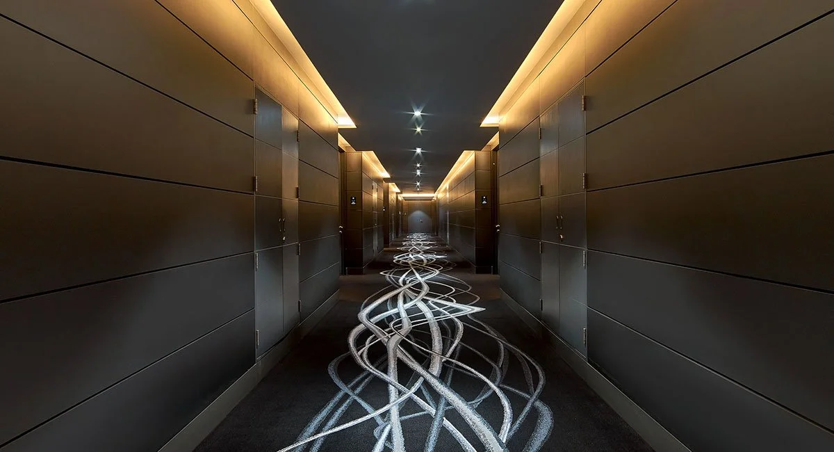 Long corridor with dark walls and ceiling, illuminated by yellow lighting along the top edges. The carpet has light-colored swirling patterns.