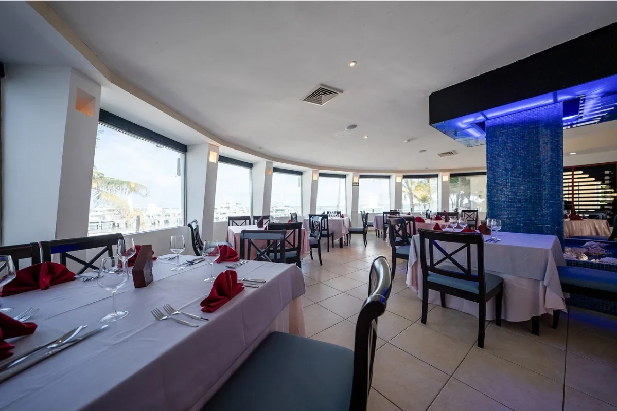 Elegant restaurant with large windows overlooking a marina, set with white tablecloths, red napkins, wine glasses, and cutlery, featuring a blue mosaic pillar and ambient lighting.