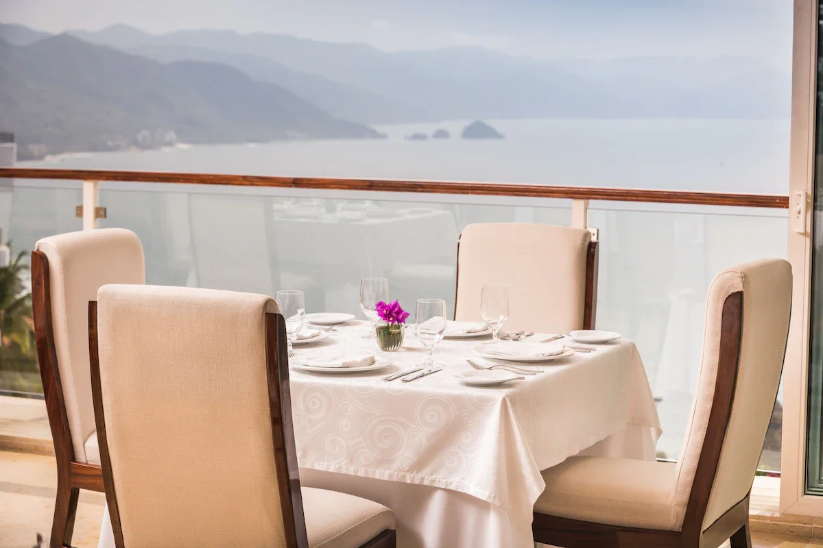 A round dining table set for a meal with a white tablecloth, plates, silverware, wine glasses, a small vase with purple flowers, and surrounded by six upholstered chairs, overlooking a balcony with a glass railing and a scenic ocean view with mountai
