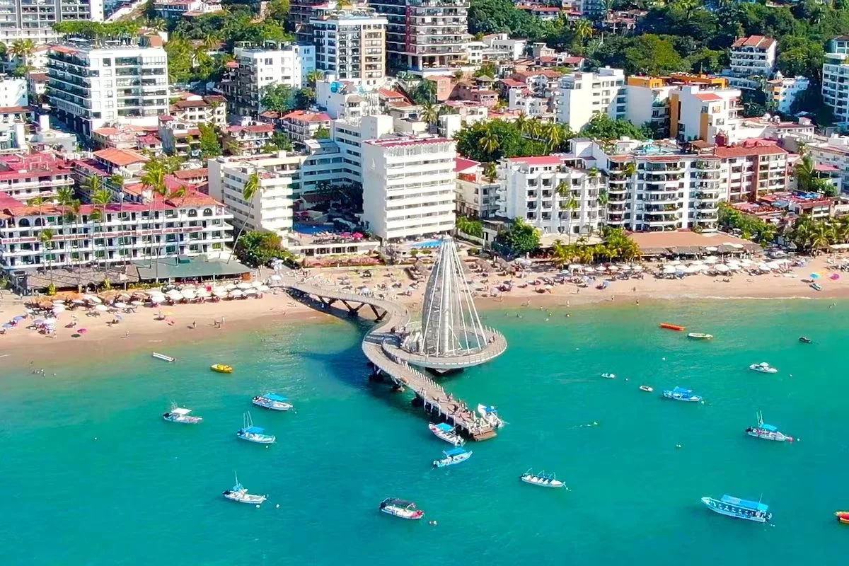 Aerial view of a coastal city with high-rise buildings, a sandy beach packed with umbrellas, and a pier with a glass-domed structure extending into turquoise water, with boats anchored nearby.