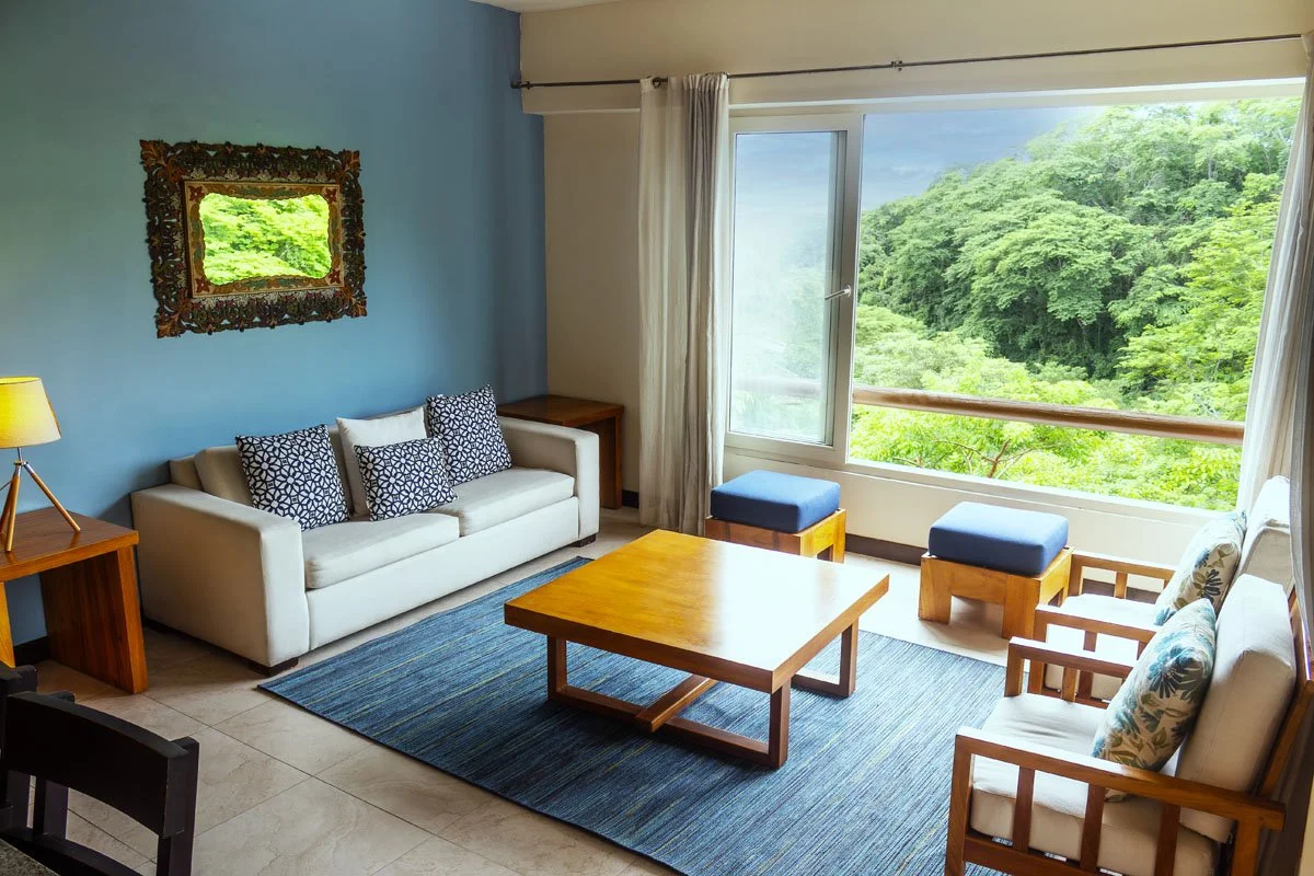 Living room with beige couch, armchair, wooden coffee table, blue ottomans, side tables, a lamp, and large window overlooking green foliage