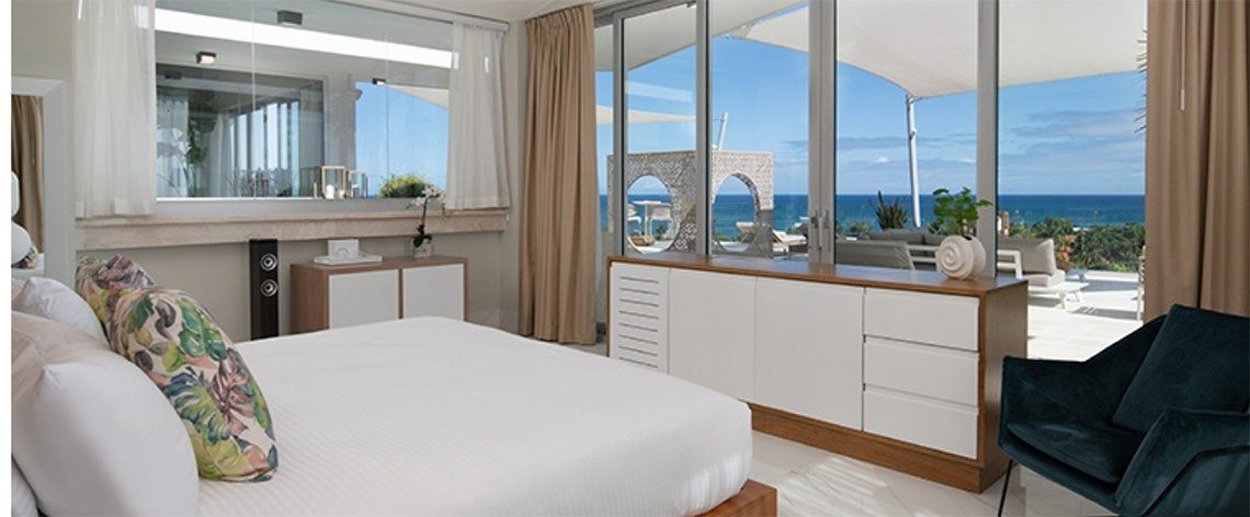 A hotel room with a large bed, colorful pillows, and a view of the ocean outside large glass doors.