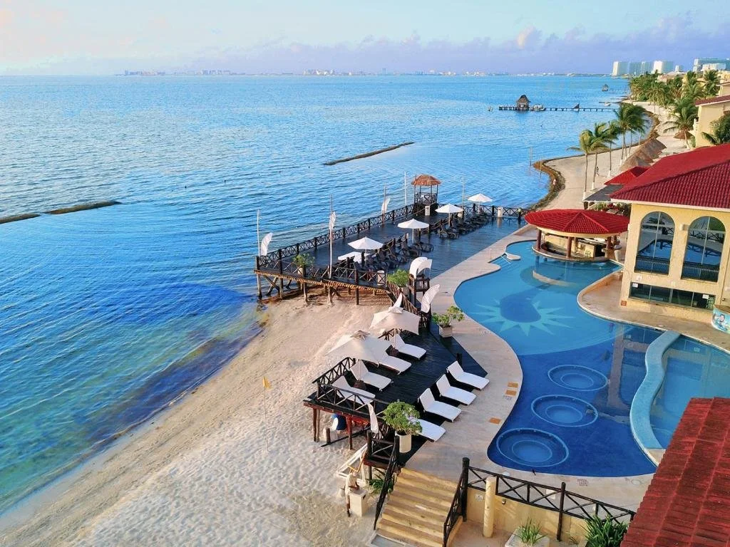 A resort with a sandy beach, a swimming pool, and a pier extending into the ocean. There are lounge chairs, umbrellas, and palm trees, with a building that has red roofs nearby.