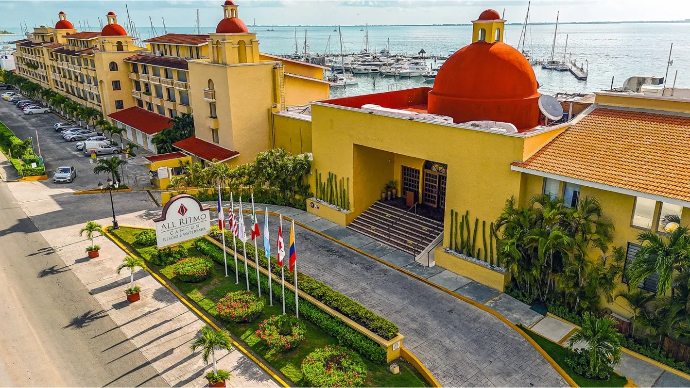 Colorful resort building with red domes, palm trees, flags, and a marina in the background at sunrise or sunset.