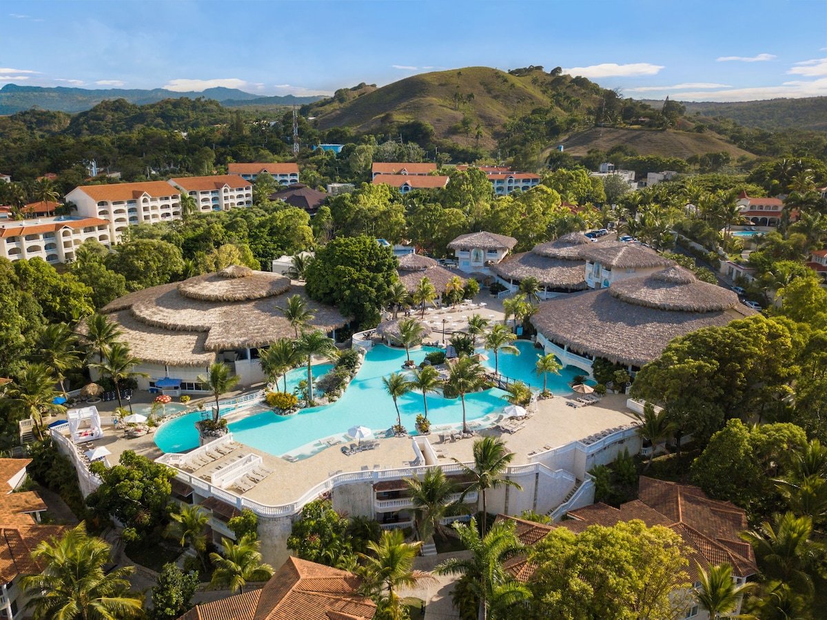 Aerial view of a tropical resort with a large swimming pool surrounded by palm trees, thatched-roof buildings, and lush greenery, with hills in the background under a blue sky.