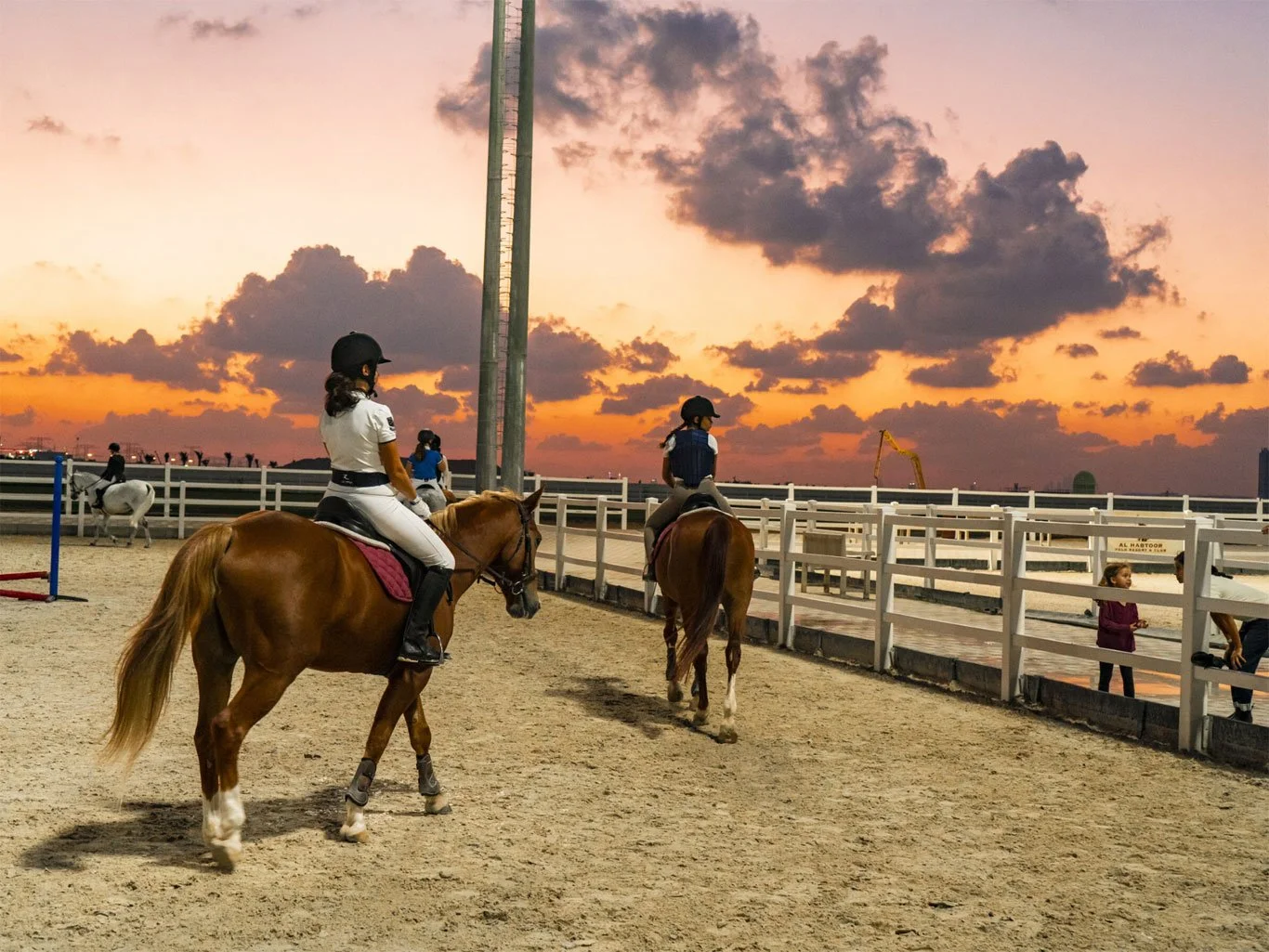 People riding horses in an outdoor arena during sunset, with a sky filled with orange and purple clouds, and a few children watching from outside the fence.