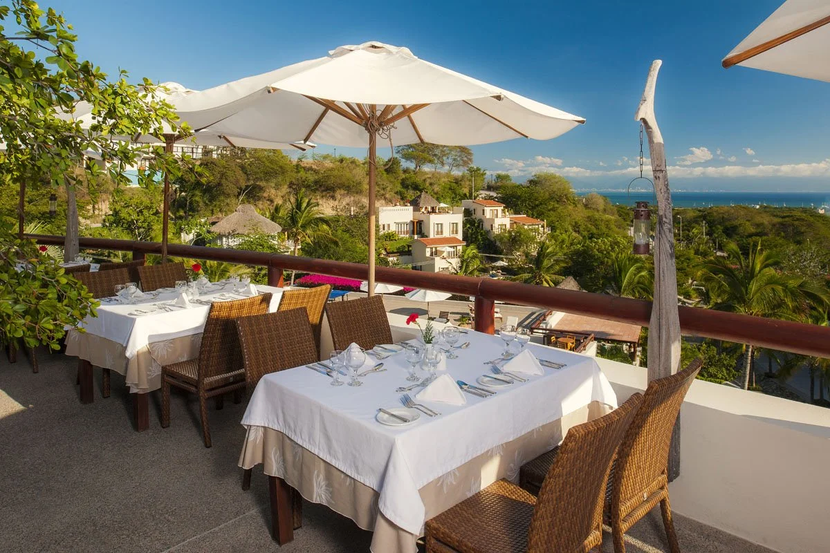 Outdoor restaurant patio with white tablecloths, wicker chairs, white umbrellas, tropical greenery, houses, and a view of the ocean in the background.