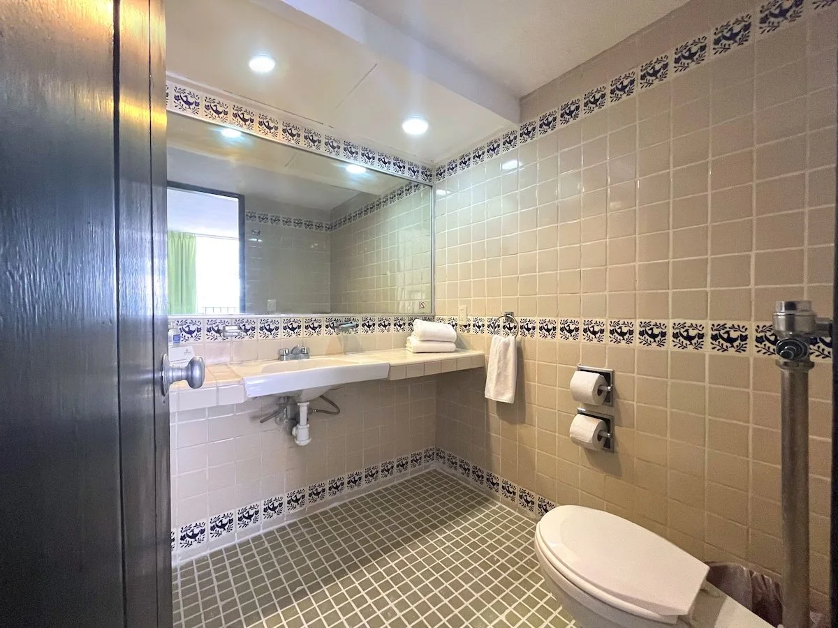 Bathroom with beige tiled walls and floor, large mirror above sink, and toilet with toilet paper rolls.