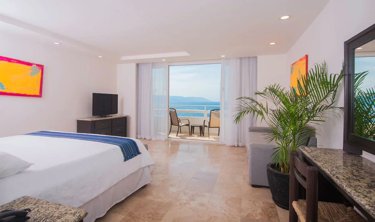 Hotel room with a large bed, a television, a potted plant, and a balcony with chairs overlooking the ocean.