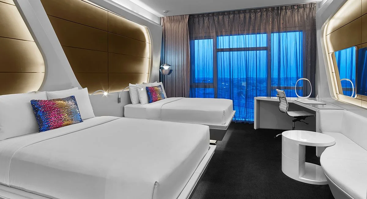 Modern hotel room with two beds, colorful pillows, a large window with blue curtains, a desk with a chair, and a circular white side table.