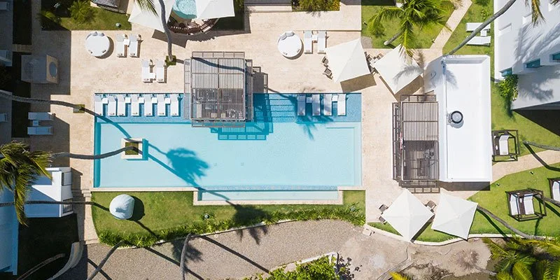 Aerial view of a luxurious pool area with lounge chairs, umbrellas, and cabanas surrounding a rectangular swimming pool in a tropical setting.