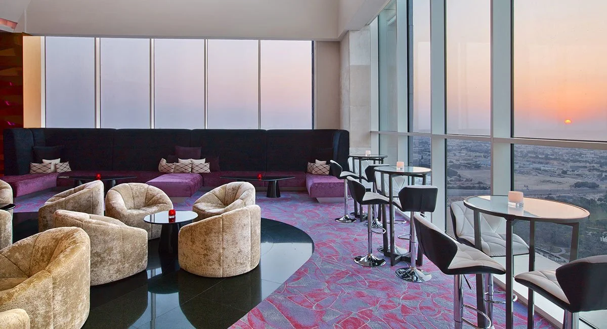 Luxury lounge with beige velvet armchairs and high tables near large floor-to-ceiling windows showing a city view at sunset.