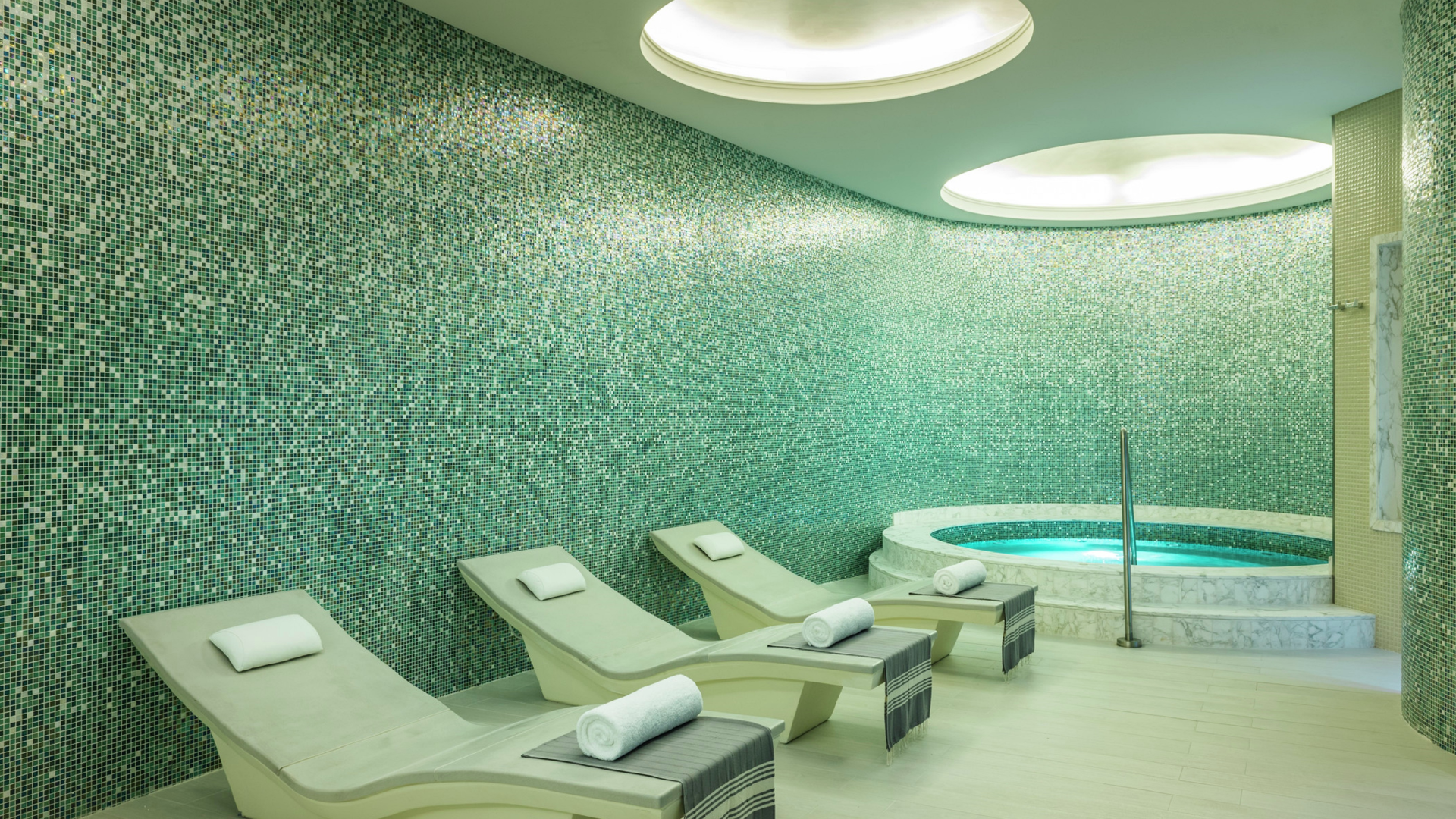 Spa room with three lounge chairs, a hot tub, and mosaic tile walls in green and blue tones, with large circular ceiling lights.