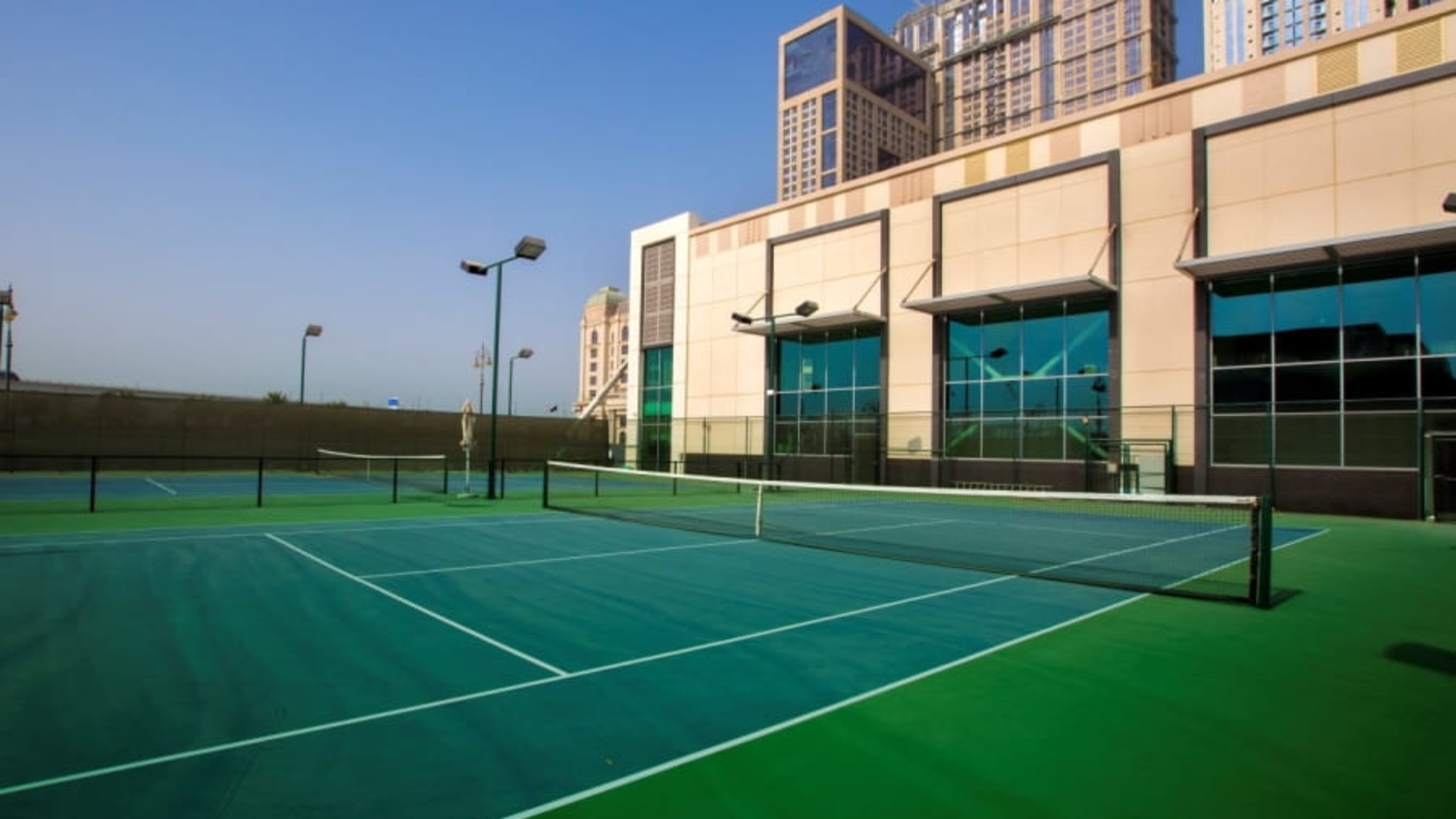 Empty outdoor tennis courts with green surface, nearby modern buildings, and clear blue sky.
