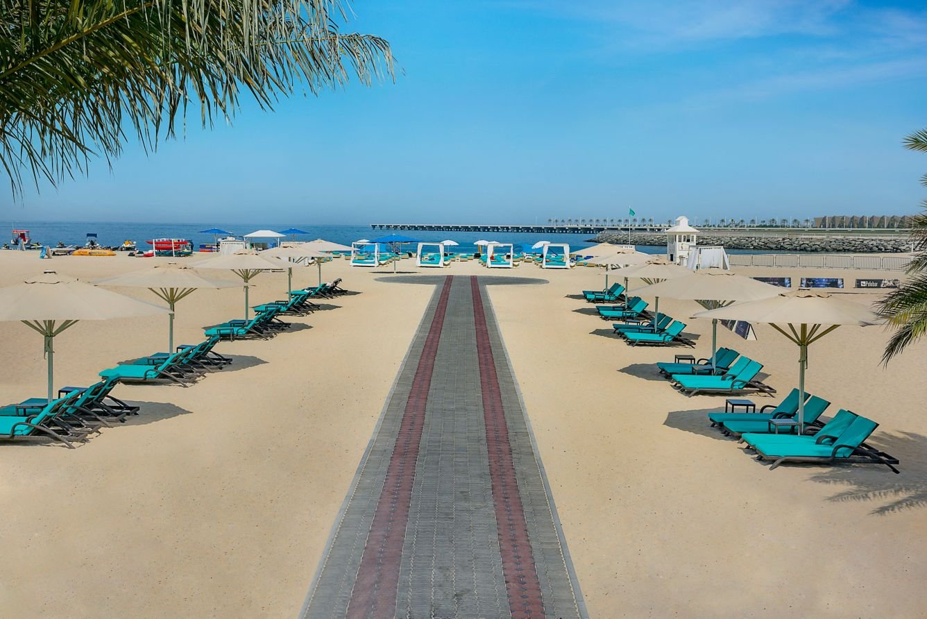 Empty beach with turquoise lounge chairs and umbrellas, walkway leading to the ocean, with lifeguard stands and a pier in the background.