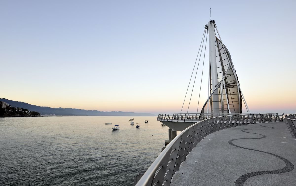 A modern waterfront promenade with a curved path and railing, a unique sail-shaped building, calm water with small boats, and distant hills at sunset.