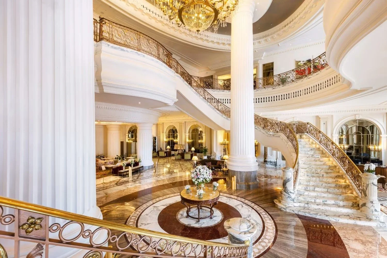 Elegant hotel lobby with white columns, marble floors, ornate staircase, chandelier, and luxurious furniture.