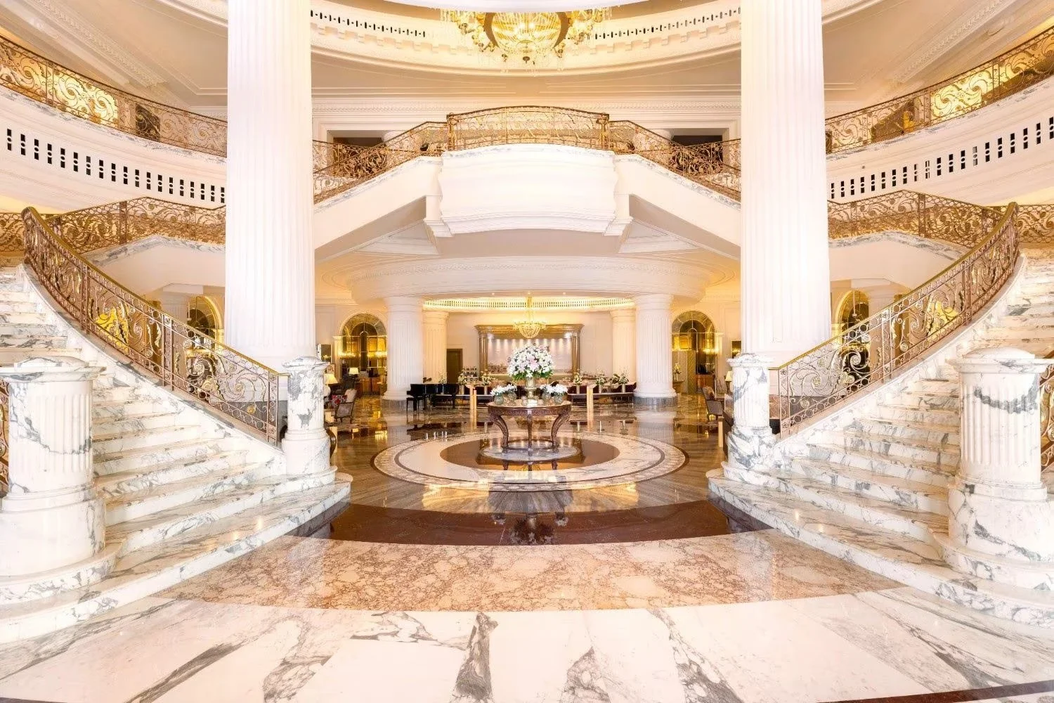Luxurious hotel lobby with marble floors, grand staircase, large columns, and ornate railings. Centered is a table with a floral arrangement, and in the background, there are seating areas and chandeliers.