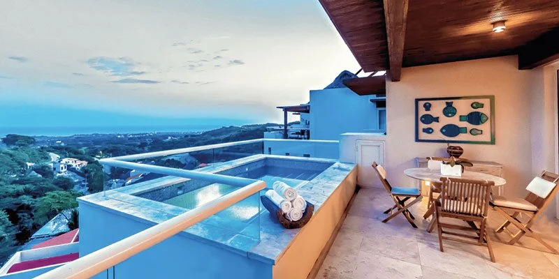 An outdoor balcony with a hot tub, seating area with wooden chairs and round table, and ocean view in the distance under a partly cloudy sky.