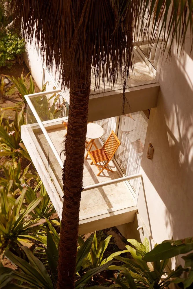 View from above of a balcony with a small white table, a wooden folding chair, and a sunlit outdoor area surrounded by plants and a tall palm tree.