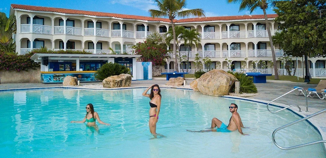 People swimming and relaxing in a hotel swimming pool, with a multi-story hotel building, palm trees, and lounge chairs in the background.